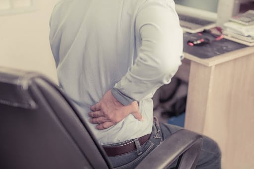 Person experiencing lower back and SI joint pain while sitting at desk, hand on lower back